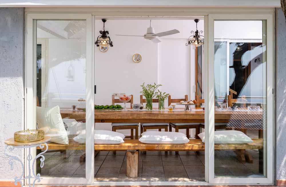 Sunlit dining area seen through sliding glass doors, featuring a rustic wooden table with benches, white cushions, glassware, and pendant lights in a minimalist coastal interior.