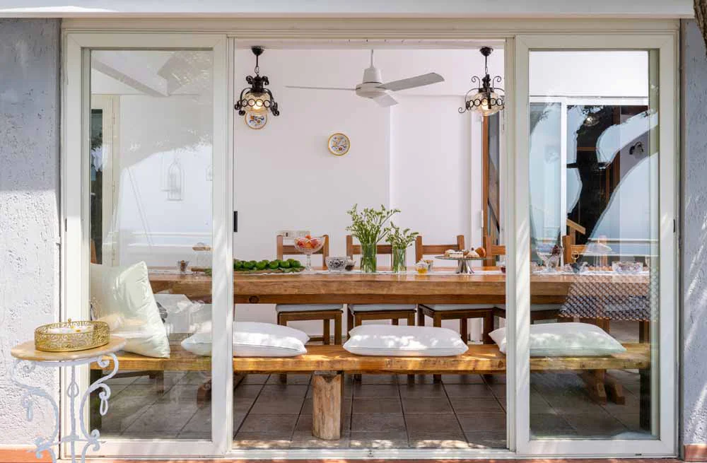 Sunlit dining area seen through sliding glass doors, featuring a rustic wooden table with benches, white cushions, glassware, and pendant lights in a minimalist coastal interior.