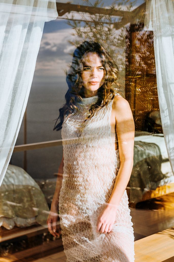 Woman in a delicate white dress framed by a glass window with warm light and reflections